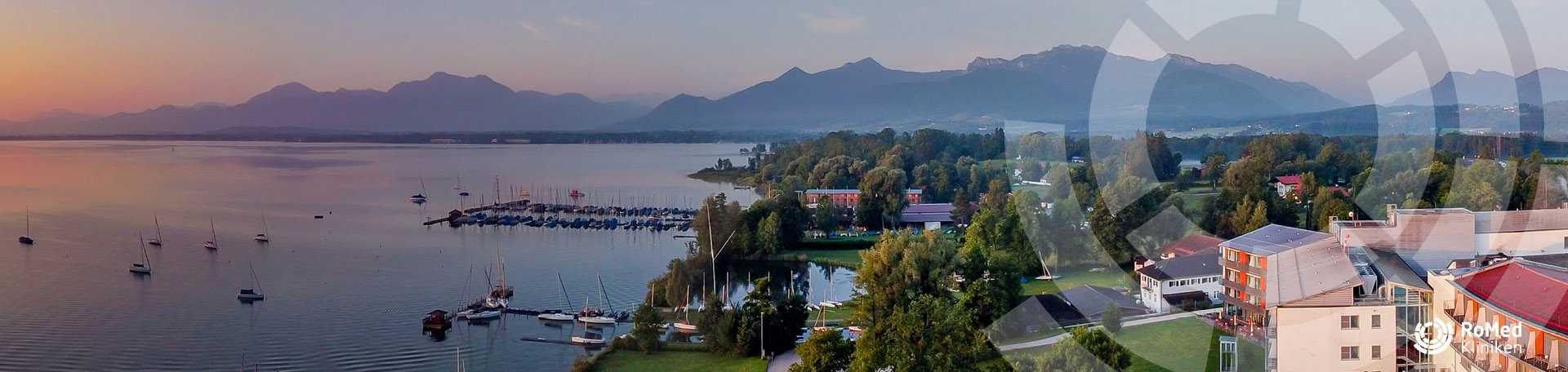 Weitwinkelaufnahme des Chiemsees mit Segelbooten und einem Yachthafen im Sonnenuntergang, links ein Uferstreifen mit der RoMed Klinik Prien und im Hintergrund eine Silhouette der Alpen.