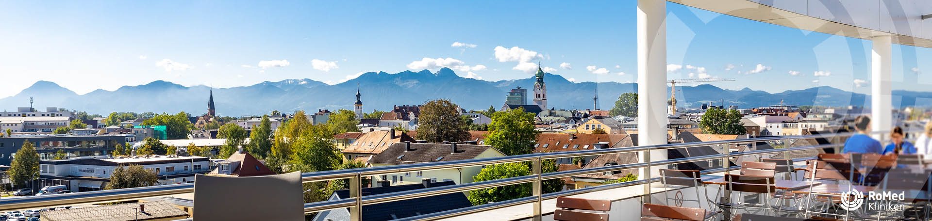 Panoramablick über die Dächer von Rosenheim mit der Gebirgskulisse der Alpen im Hintergrund, von der  Kantinenterrasse mit Tischen und Stühlen im Vordergrund.