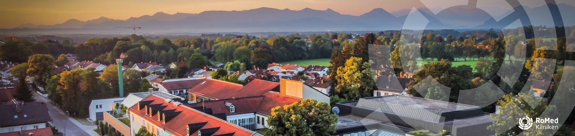 Dächer der RoMed Klinik Bad Aibling im Vordergrund, umgeben von einem Wohngebiet und grünen Bäumen, mit einer weiten Bergsilhouette im Hintergrund bei Sonnenuntergang.