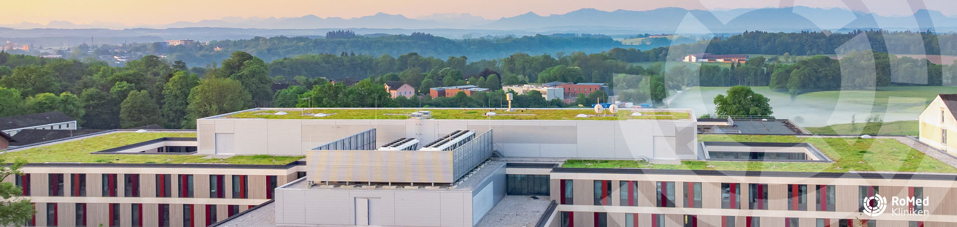 Dachlandschaft der RoMed Klinik Wasserburg mit begrünten Flachdächern und Lüftungseinheiten, im Hintergrund eine grüne Hügellandschaft mit Bergsilhouette.