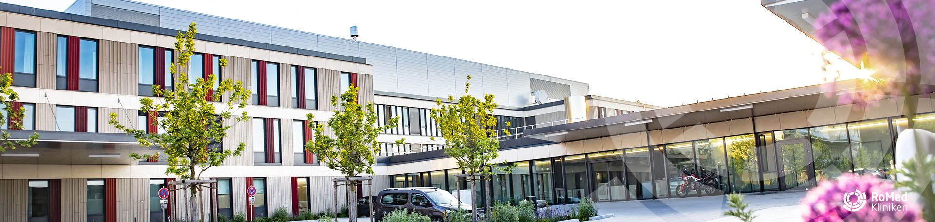 Außenansicht der modernen RoMed Klinik Wasserburg mit heller Fassade, vertikalen roten Akzenten an den Fenstern und jungen Bäumen im Vorplatzbereich.